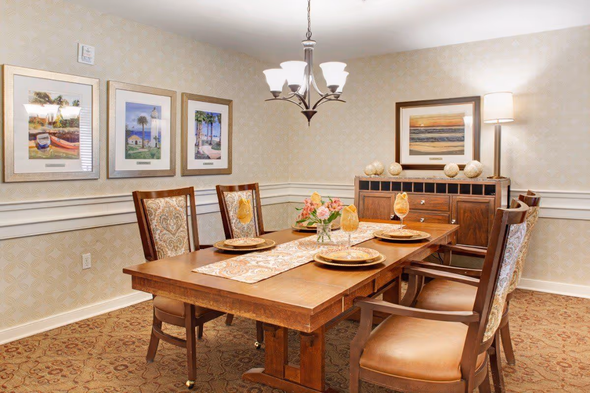 Dining room with a wooden table set with plates and glasses, upholstered chairs, framed artwork, chandelier, and a sideboard.