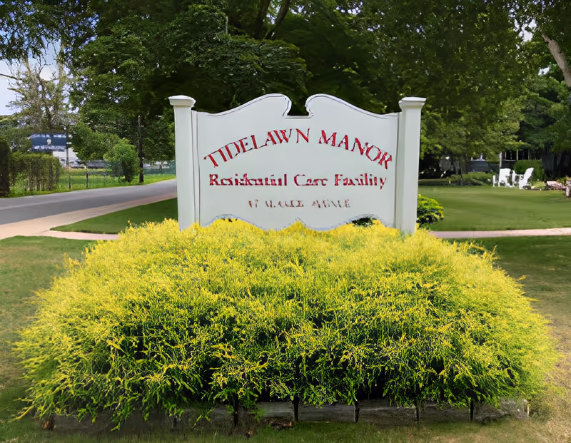 A white entrance sign for 'Tidelawn Manor' set among trimmed shrubs on a lawn with trees and a road behind it.