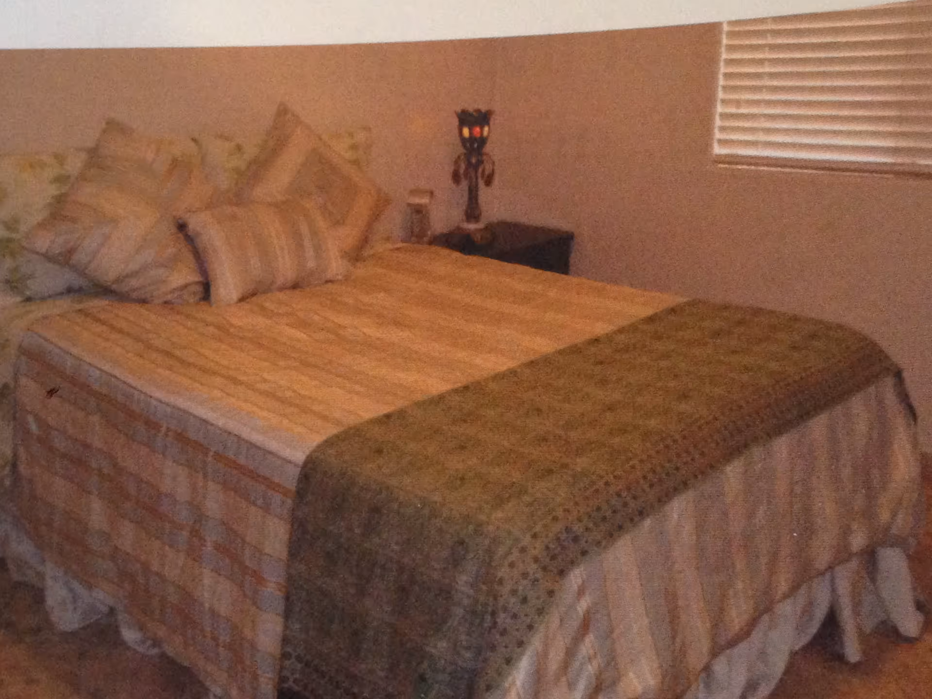 A neatly made bed with striped bedding, decorative pillows, a green throw, and a nightstand under a window with blinds.