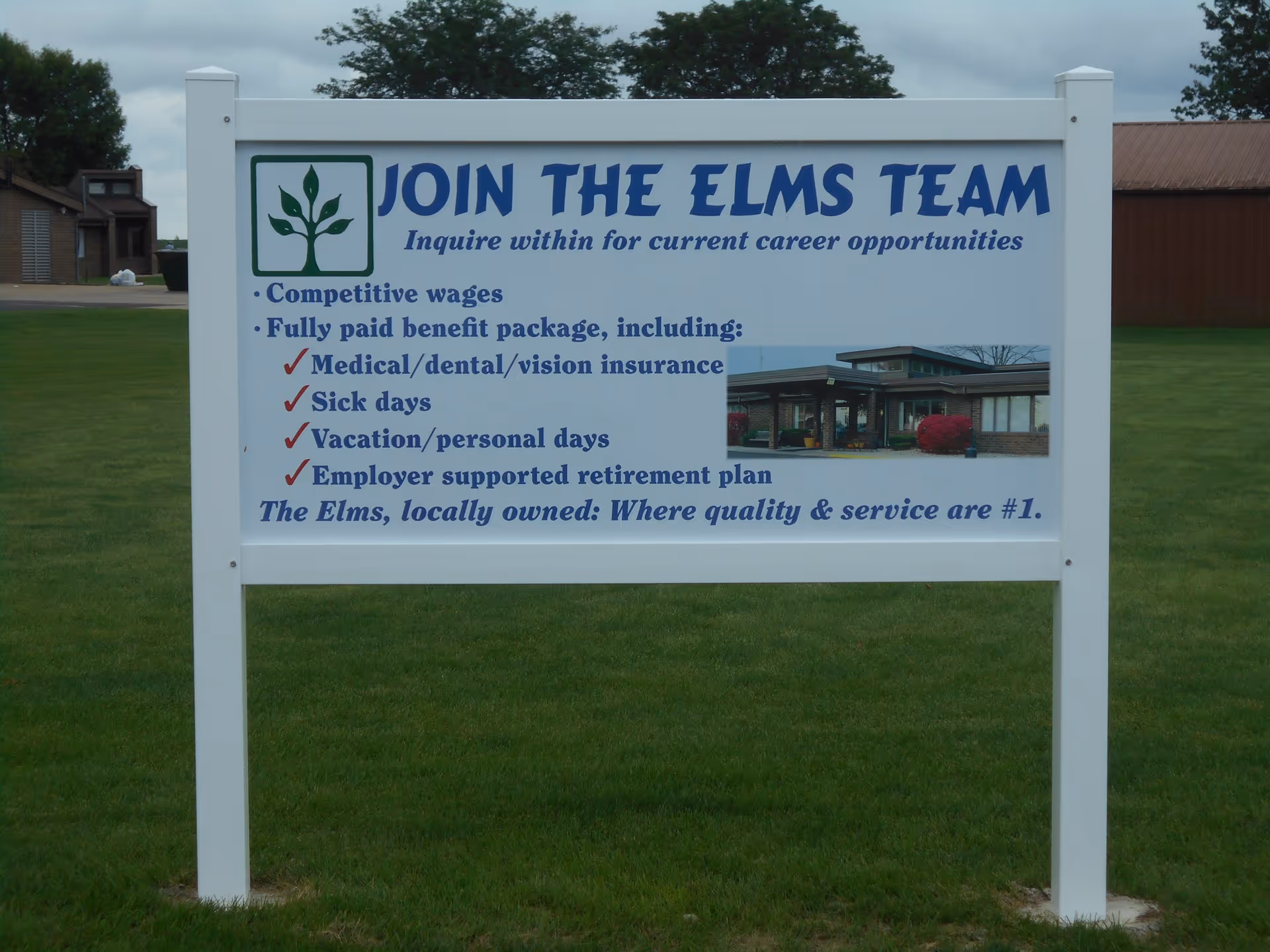 A white sign on a grassy area with the heading 'JOIN THE ELMS TEAM' inviting inquiries for current career opportunities. The sign lists benefits including competitive wages, fully paid benefit package with medical/dental/vision insurance, sick days, vacation/personal days, and employer supported retirement plan. There is a small photo of a building on the sign and a logo of a green tree. The bottom of the sign reads 'The Elms, locally owned: Where quality & service are #1.'