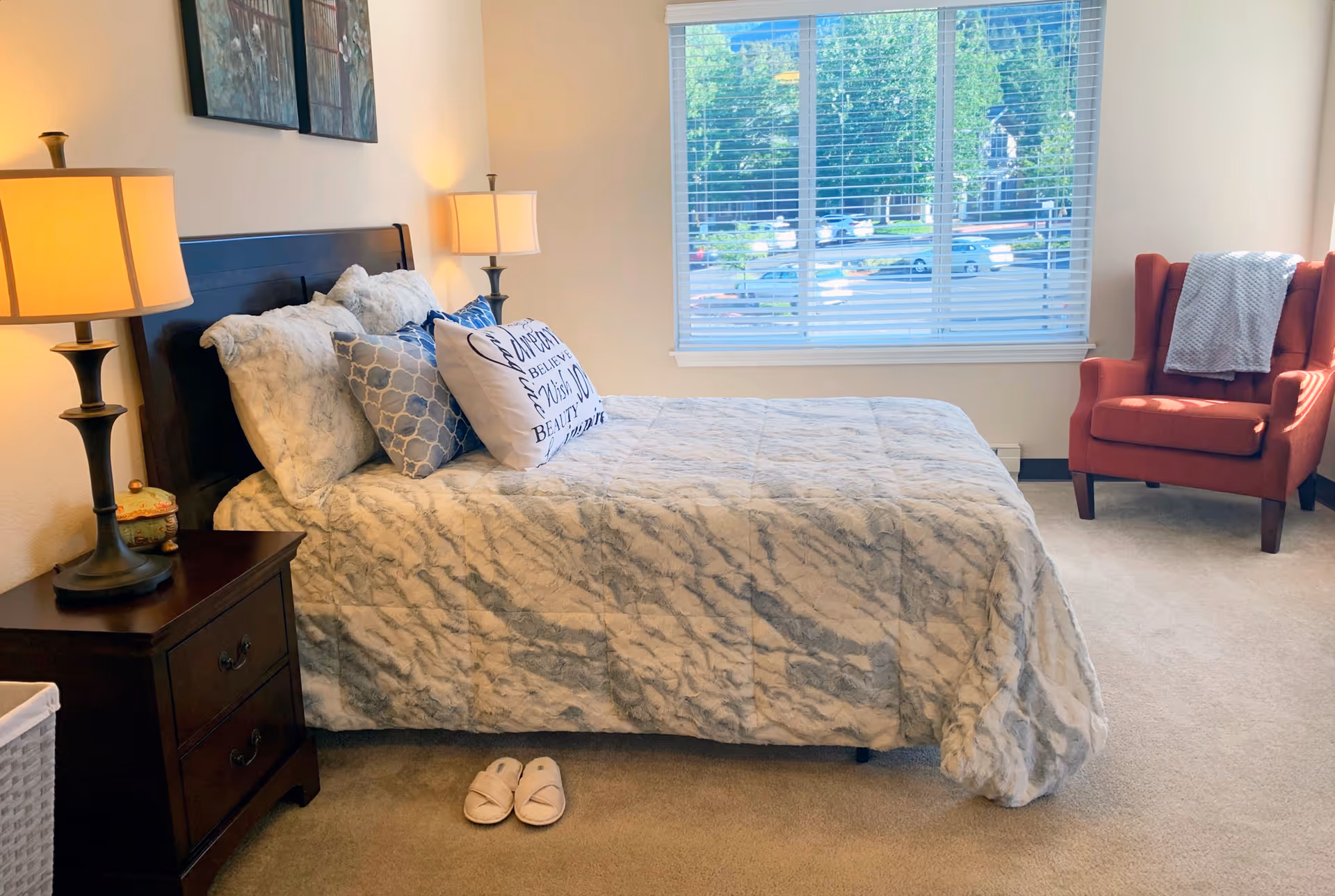 A cozy bedroom with a neatly made bed covered in a light gray and white patterned comforter, several decorative pillows, two bedside tables with lamps, a red armchair with a gray blanket draped over it, and a large window with blinds showing a view of trees and parked cars outside.