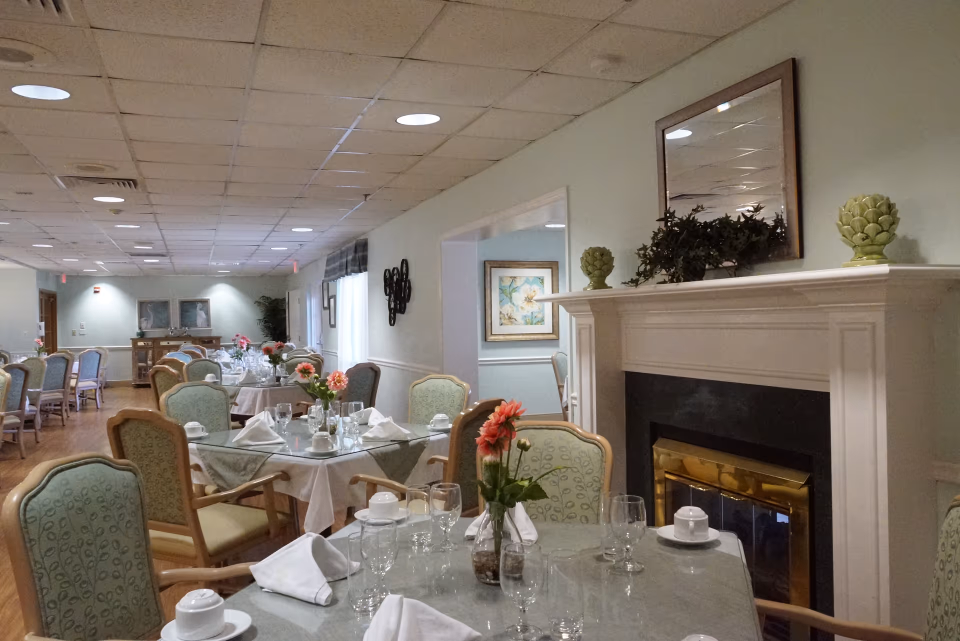 A dining room in a senior living facility with multiple tables set with white napkins, glassware, and small flower arrangements. The room features light green upholstered chairs, a fireplace with decorative items on the mantel, and soft lighting from ceiling fixtures.