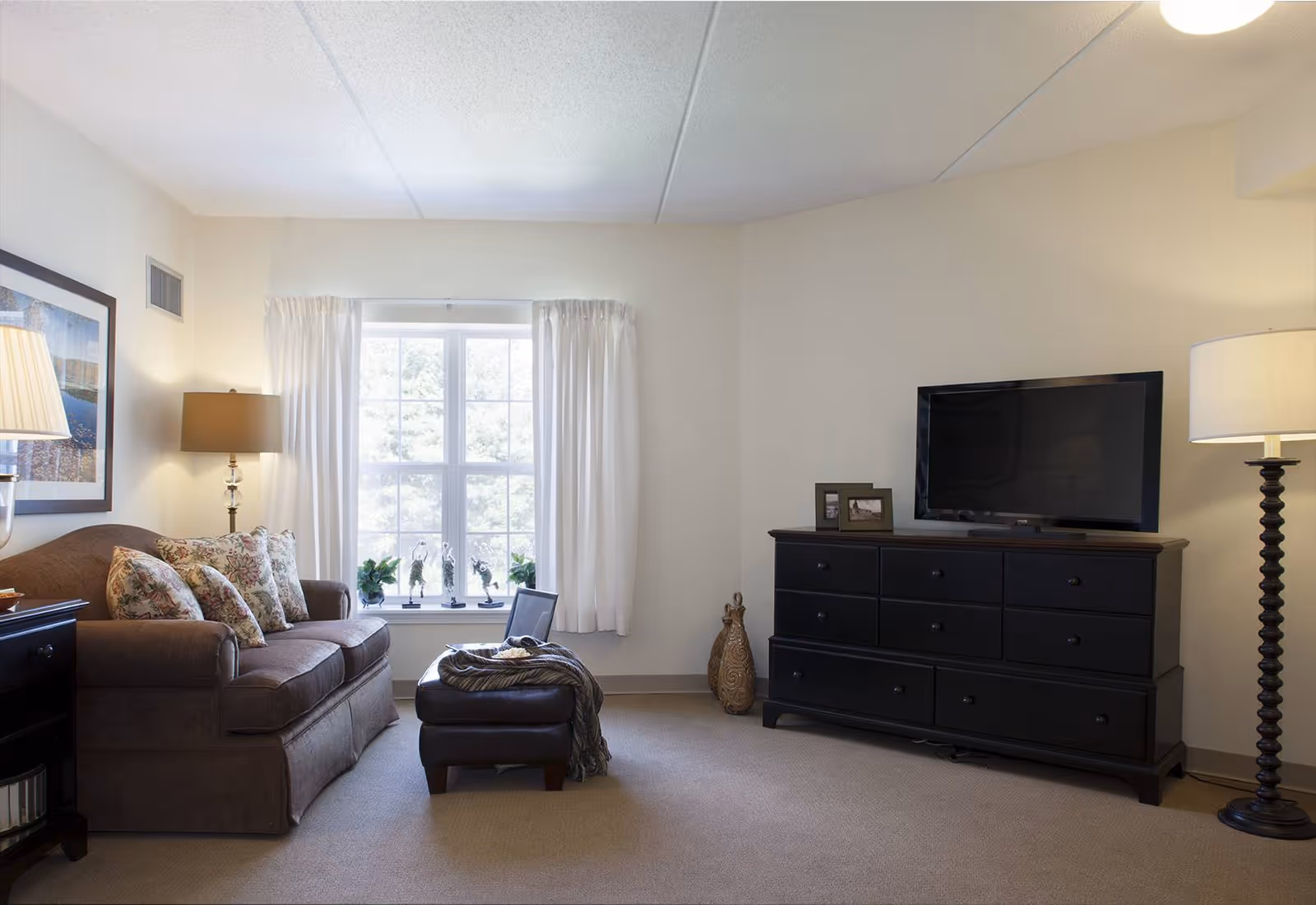 A cozy living room with a sofa and patterned pillows, an ottoman, a dresser with a television, floor lamps, and a window with white curtains.