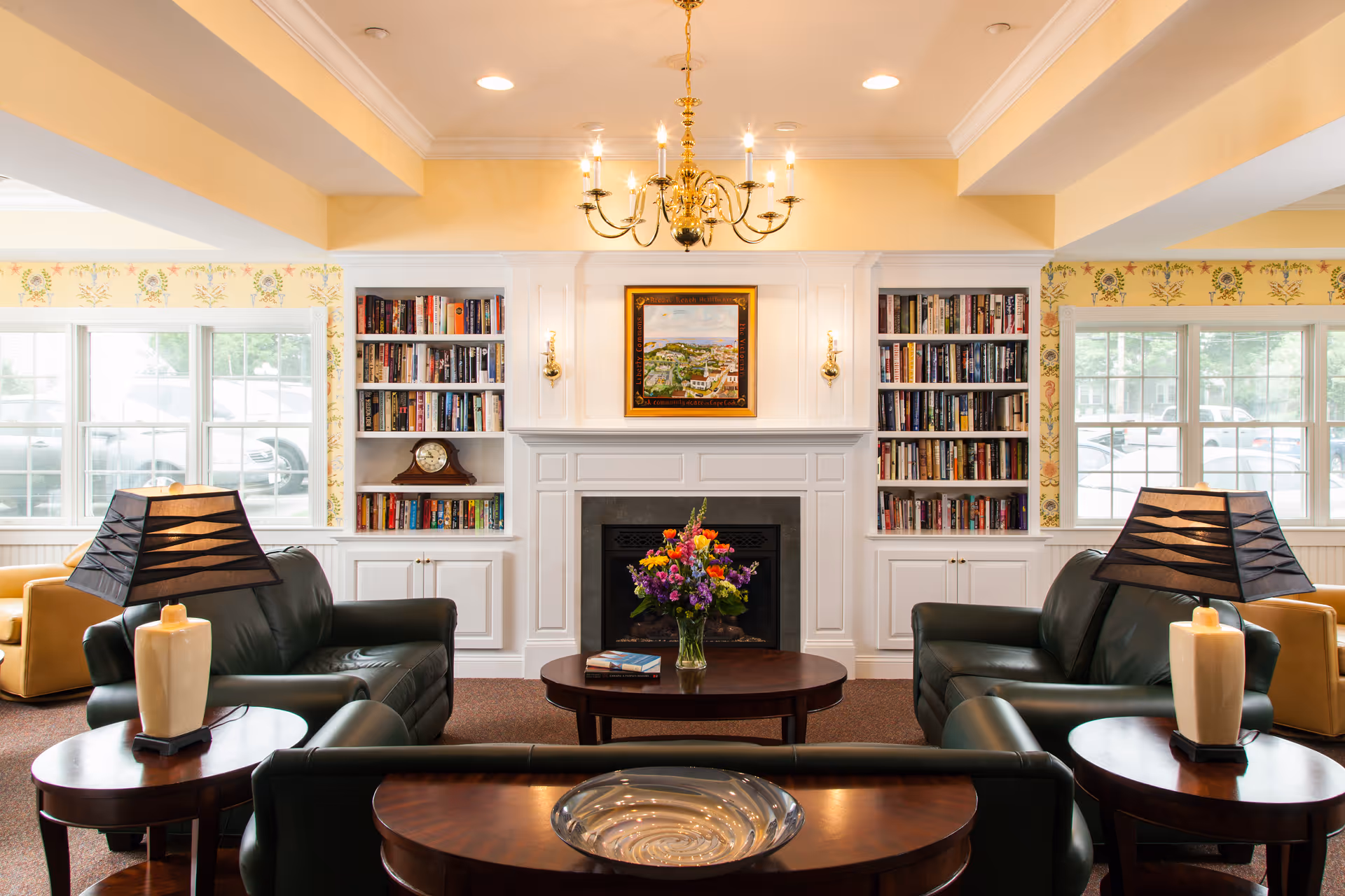A cozy living room with two dark green leather armchairs and a matching sofa arranged around a wooden coffee table with a vase of colorful flowers. The room features a white fireplace with built-in bookshelves on either side filled with books. Above the fireplace hangs a framed painting, and a gold chandelier is suspended from the ceiling. Two table lamps with black shades sit on wooden side tables next to the armchairs. Large windows on both sides let in natural light, and the walls have a floral wallpaper border.