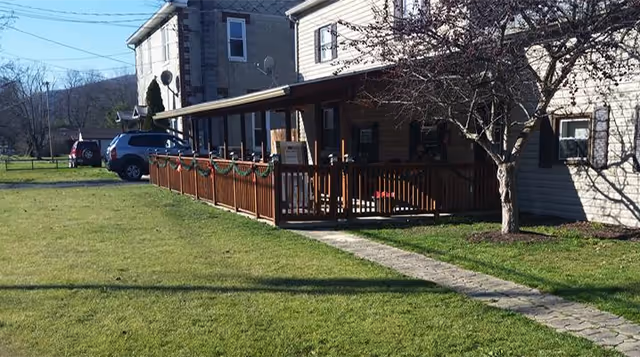 Single-story building with a covered front porch and wooden railing, a stone walkway across a grassy lawn, and cars parked to the side.