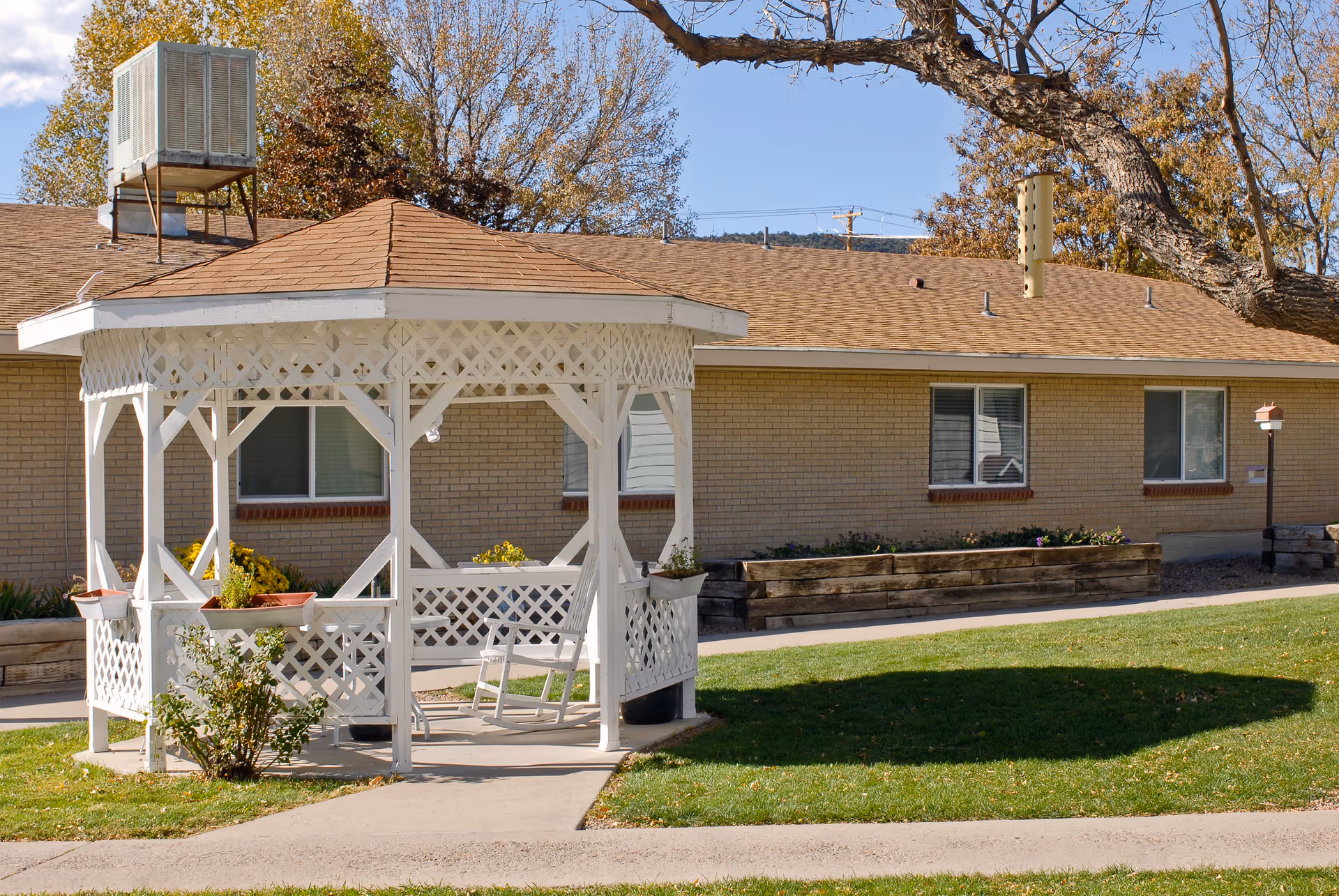 A white wooden gazebo with lattice design stands on a concrete path surrounded by green grass. Behind the gazebo is a single-story brick building with several windows and a brown shingled roof. There are some trees with autumn-colored leaves in the background under a clear blue sky.