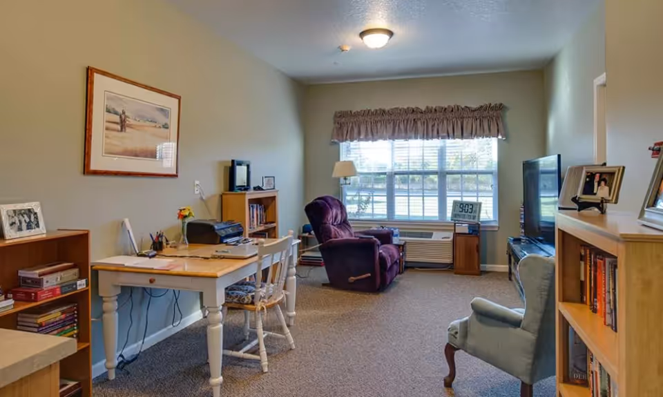 A cozy living room area in a senior living facility with a purple recliner chair, a light blue armchair, a wooden desk with a chair, bookshelves filled with books, a large window with blinds and a valance, a TV on a stand, and framed pictures on the walls and shelves.
