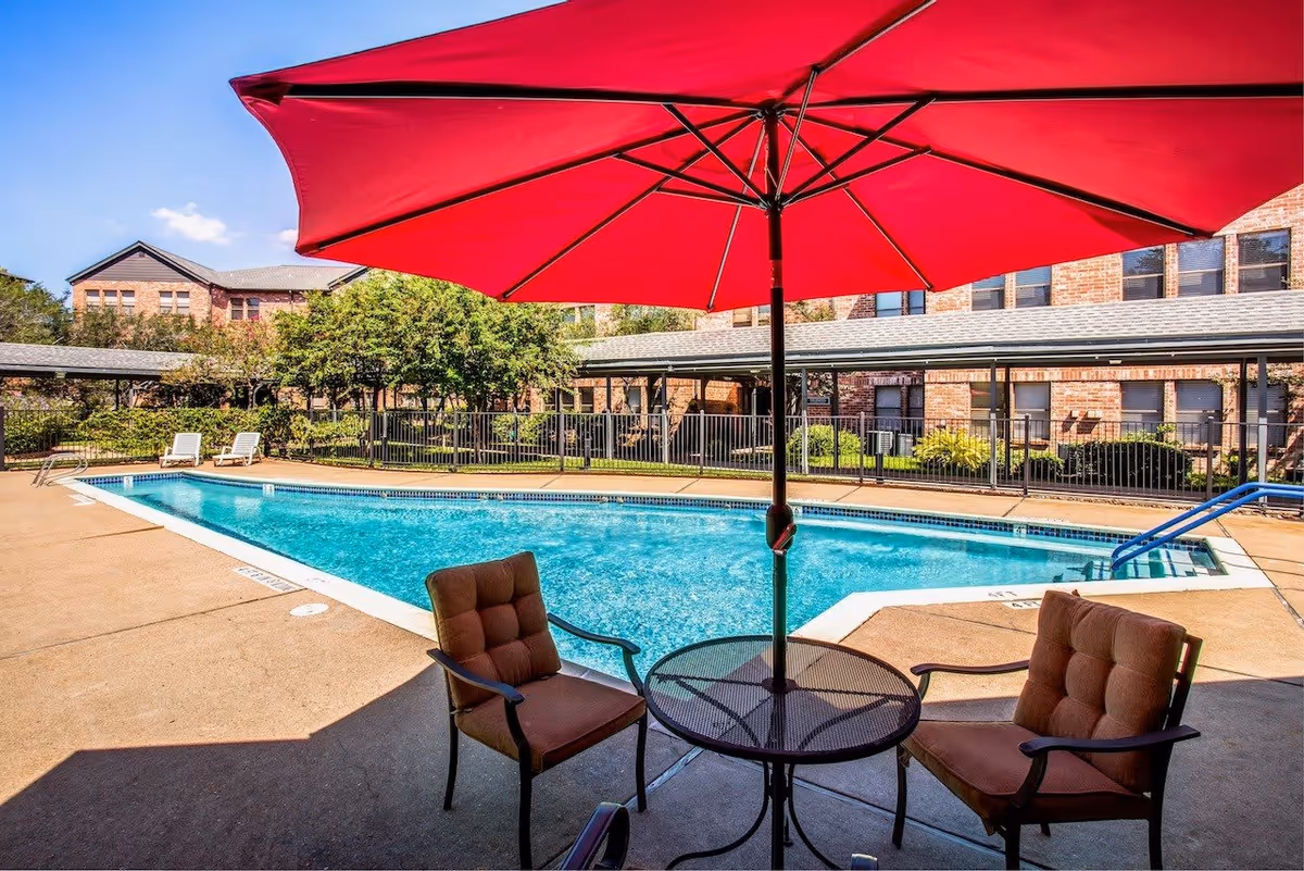 Outdoor swimming pool area with clear blue water, surrounded by a concrete deck. There are two cushioned chairs and a round metal table under a large red umbrella in the foreground. In the background, there are brick buildings, greenery, and two white lounge chairs near the pool.