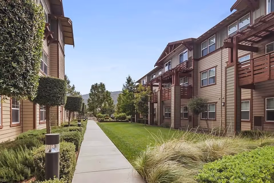 A landscaped outdoor walkway between two multi-story residential buildings with neatly trimmed bushes, trees, and grass under a clear blue sky.