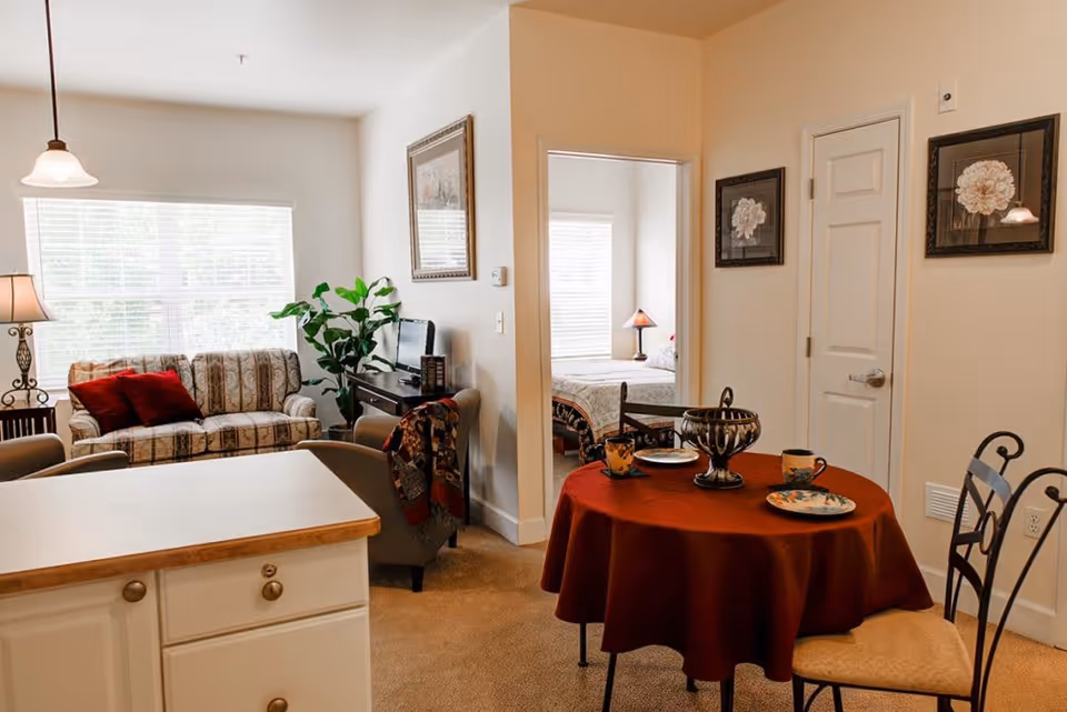 Interior view of a senior living facility showing a small dining area with a round table covered with a red tablecloth, set with plates and mugs. In the background, there is a living room with a patterned sofa, armchair, side table with a lamp, and a plant near a window with blinds. A bedroom is visible through an open doorway, featuring a bed with a lamp on the nightstand. The walls are decorated with framed floral artwork.
