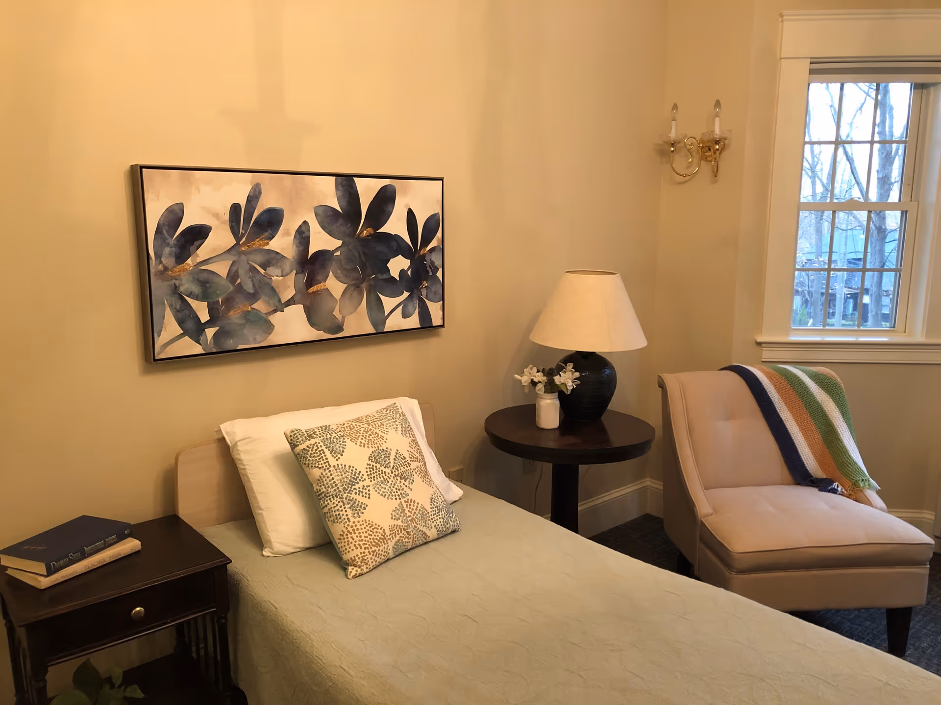 A cozy bedroom with a single bed covered in a light green quilt and two pillows, one white and one patterned. Above the bed hangs a framed artwork featuring large blue and green leaves. To the left of the bed is a dark wooden nightstand with two books on it. To the right is a round wooden side table with a black lamp and a small vase with white flowers. Next to the table is a beige upholstered chair with a colorful striped blanket draped over the back. A window with white trim lets in natural light, and a brass wall sconce is mounted on the wall beside it.