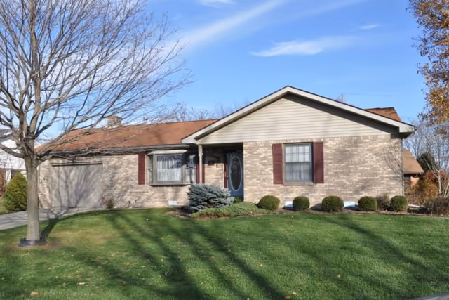 Single-story brick house with a brown shingled roof, a front door with an oval glass panel, two windows with maroon shutters, a small landscaped area with bushes and a tree without leaves, and a well-maintained green lawn under a clear blue sky.