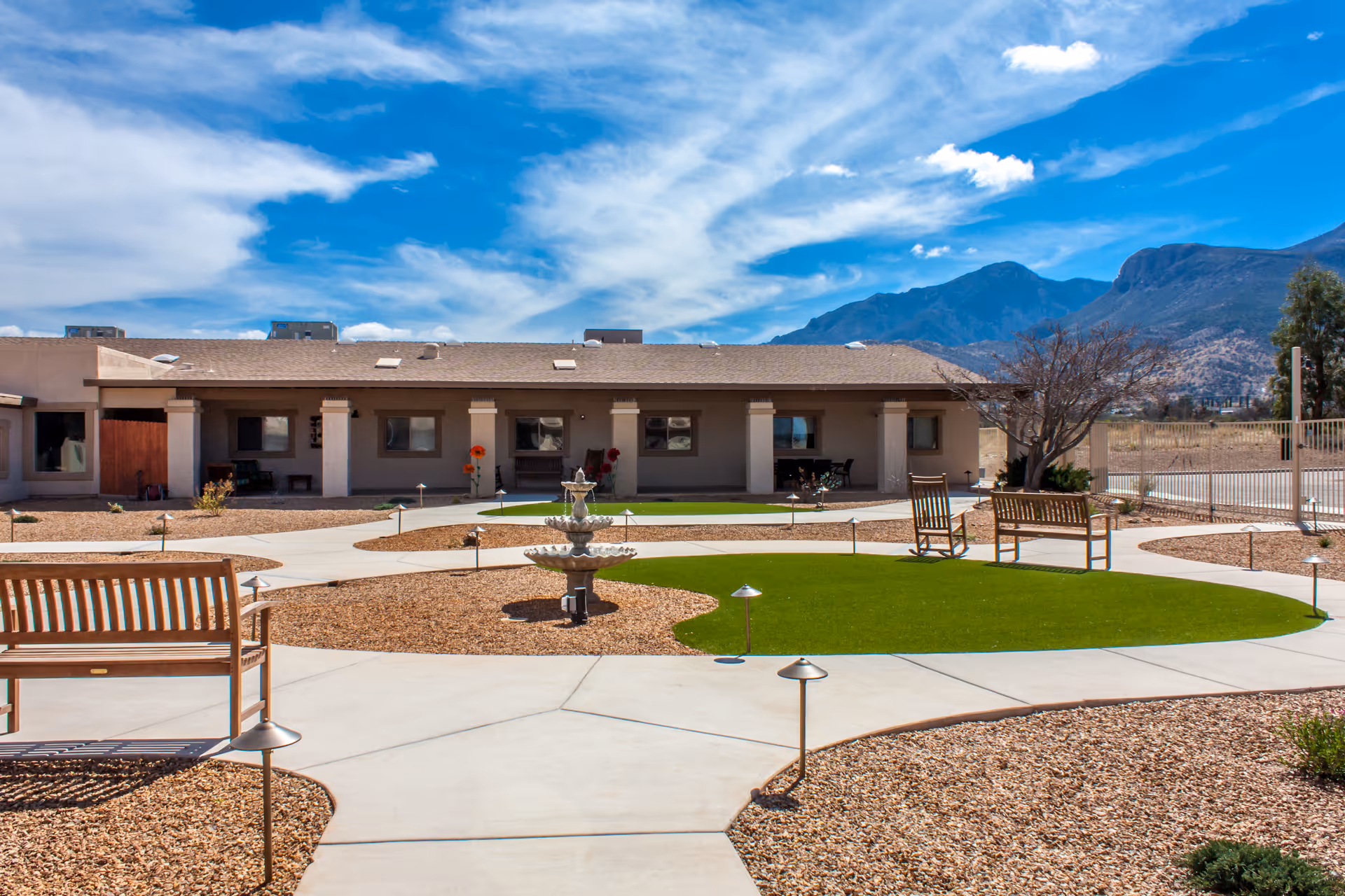 Outdoor courtyard area of Via Elegante Assisted Living Sierra Vista Canyons featuring a paved walkway, green artificial turf patches, wooden benches, a multi-tiered fountain, and a backdrop of mountains under a partly cloudy blue sky.