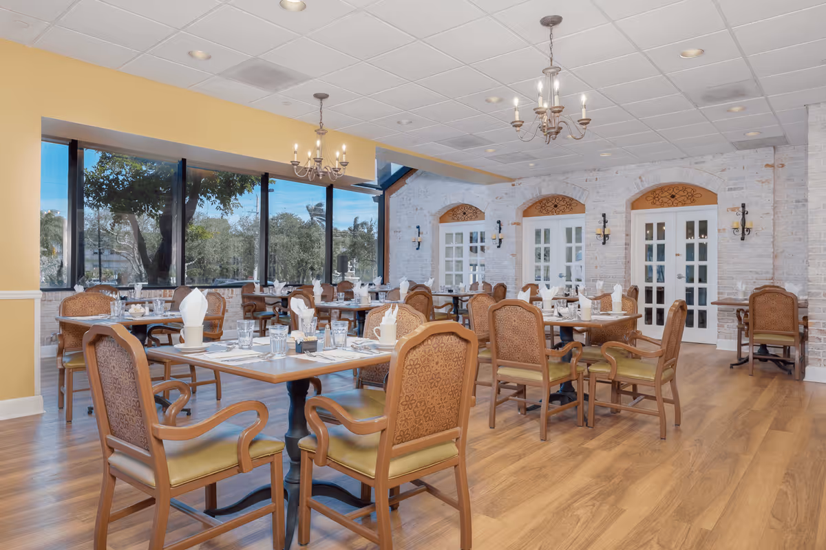 Bright, spacious dining room with multiple set tables and wooden chairs, chandeliers, large windows and French doors.