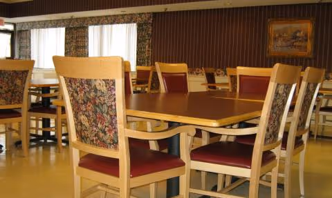 A dining room with wooden tables and chairs featuring floral patterned backs and red cushioned seats. The room has large windows with sheer white curtains, a dark wood-paneled wall, and a framed painting hanging on the wall.