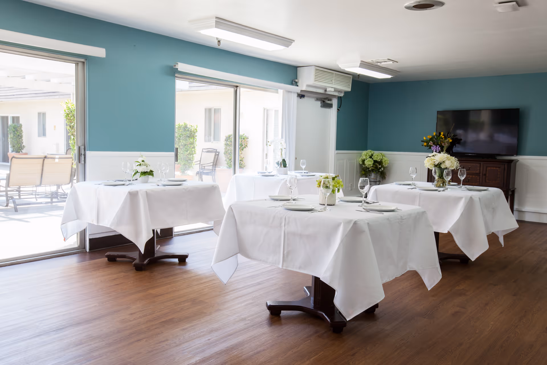A bright dining room with four tables covered in white tablecloths, each set with plates, glasses, and floral centerpieces. The room has teal walls with white wainscoting, wood flooring, large sliding glass doors leading to an outdoor patio, and a flat-screen TV mounted on the wall.