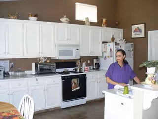 A woman in a purple shirt stands in a kitchen with white cabinets and appliances. The kitchen includes a stove, microwave, toaster oven, and refrigerator. There are decorative items on top of the cabinets and a small island with a plant and a green cup. A dining table with a floral tablecloth and white chairs is partially visible.