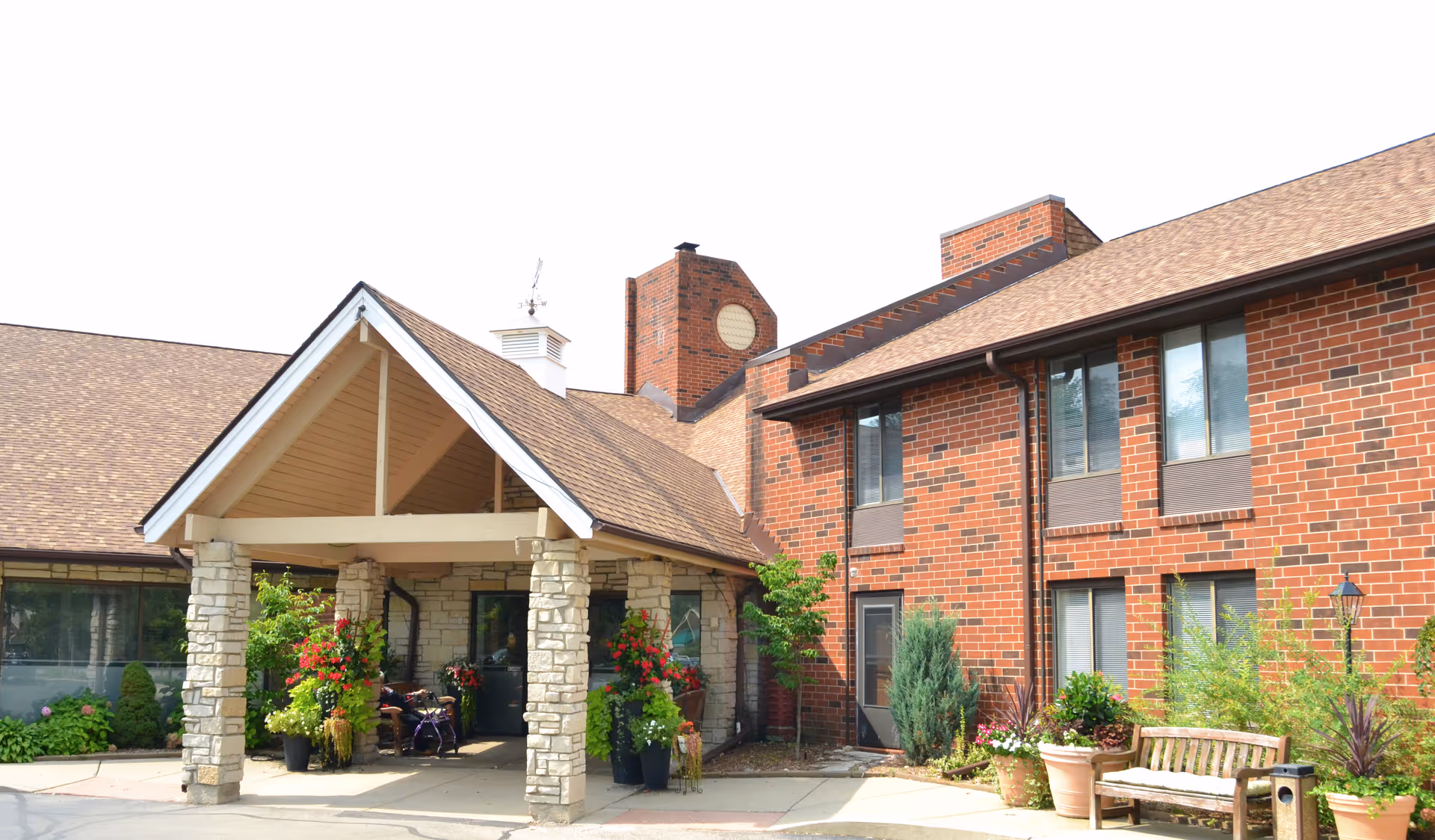 Entrance to a senior living facility with a covered porch supported by stone pillars, surrounded by potted plants and flowers. The building features red brick walls and multiple windows. There is a wooden bench and additional greenery near the entrance.