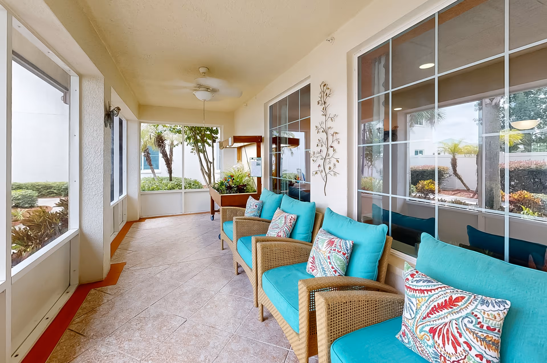 A bright enclosed patio area with large windows showing outdoor greenery. The space features a row of wicker chairs with turquoise cushions and colorful patterned pillows. There is a ceiling fan above and a decorative metal wall art piece on the wall. A planter with green plants is visible at the far end near the windows.