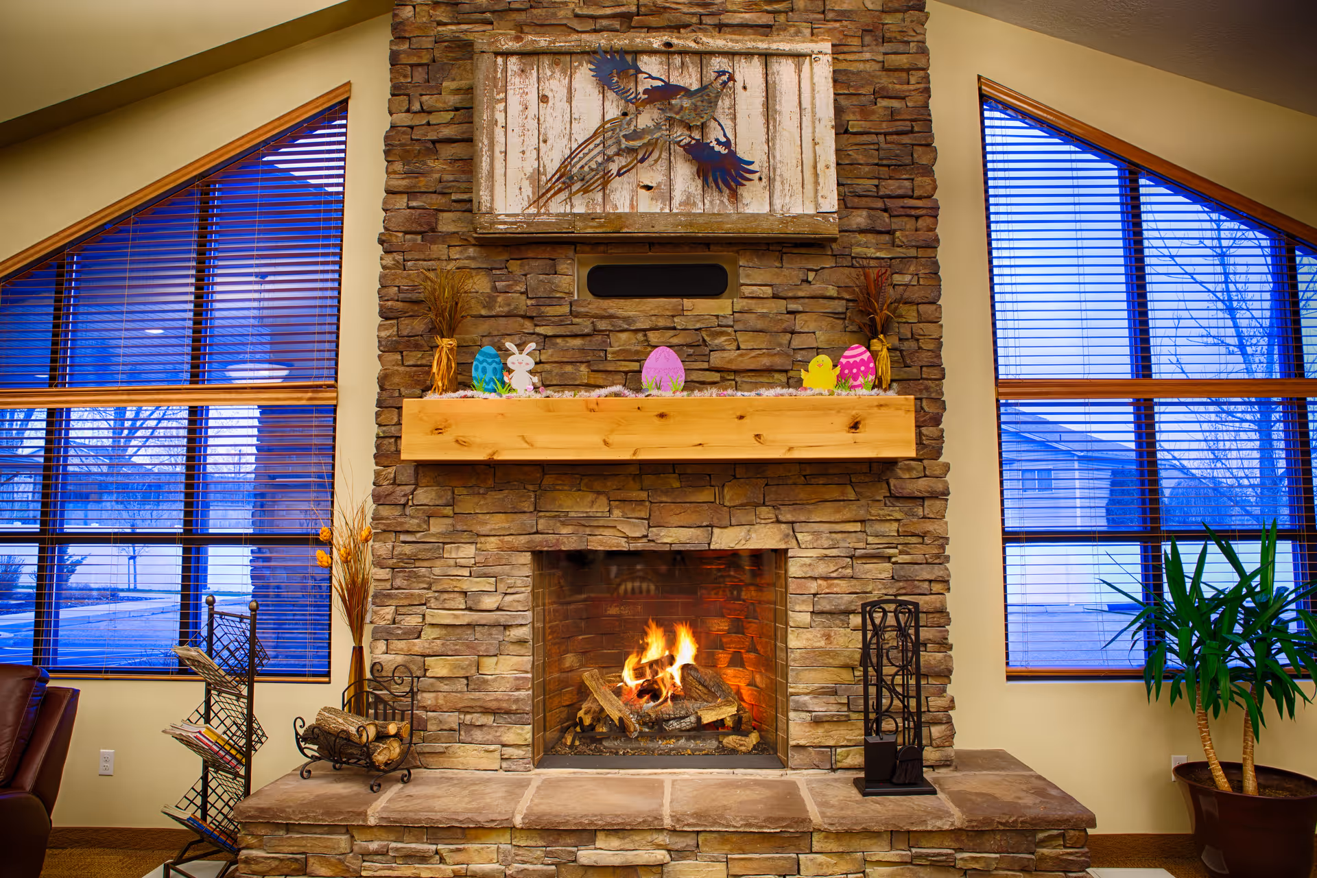 A cozy stone fireplace with a wooden mantel decorated with colorful Easter-themed ornaments including eggs, a bunny, and a chick. Above the mantel is a rustic wooden panel featuring a metal bird sculpture. On either side of the fireplace are large windows with blue blinds, and there is a potted plant on the right side and a magazine rack on the left.