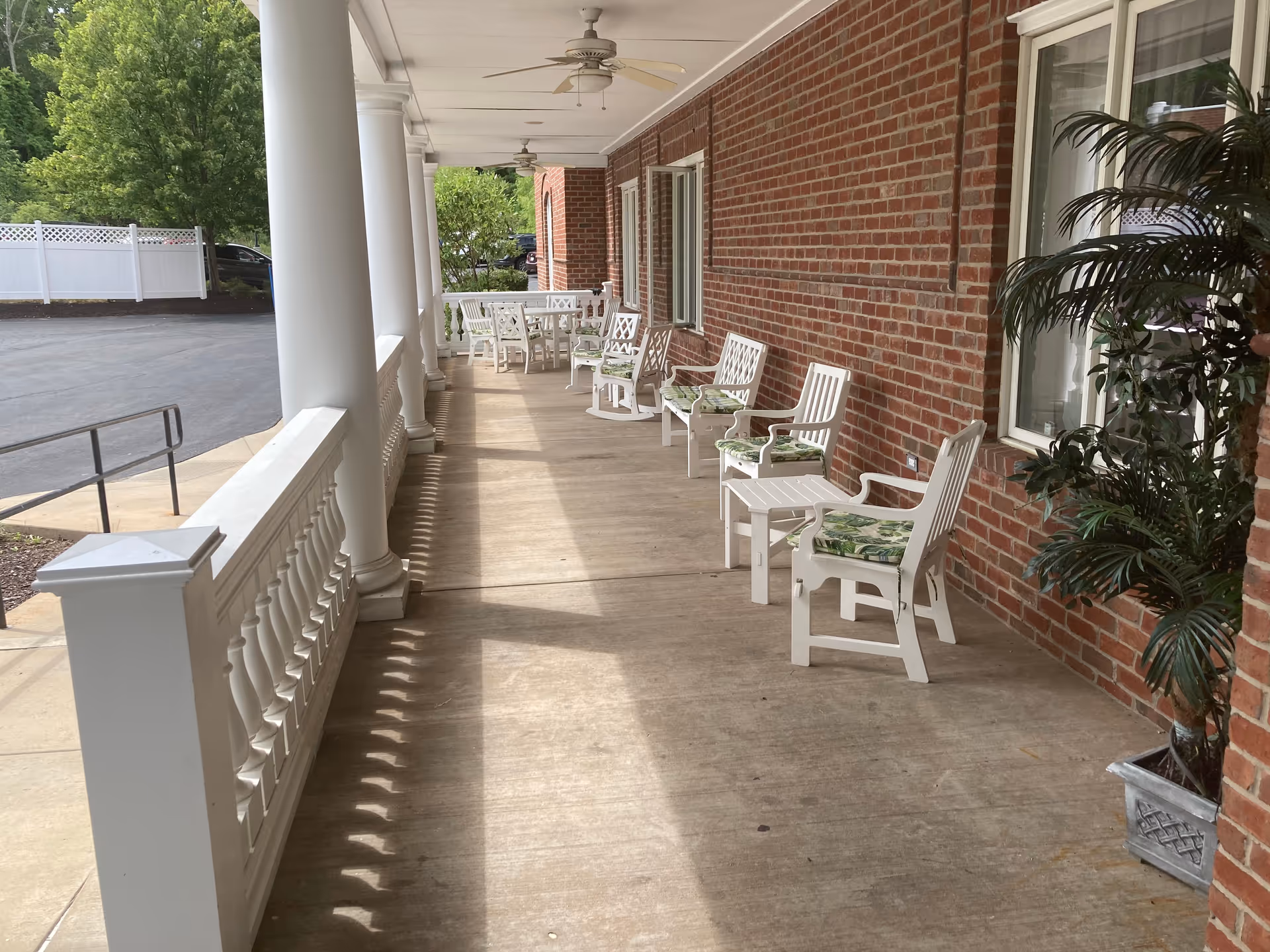 A covered outdoor porch area with white columns and a brick wall. Several white chairs with green patterned cushions and small white tables are arranged along the wall. Ceiling fans are mounted on the porch ceiling, and a potted plant is visible near the windows. The porch overlooks a paved area with greenery and a white fence in the background.