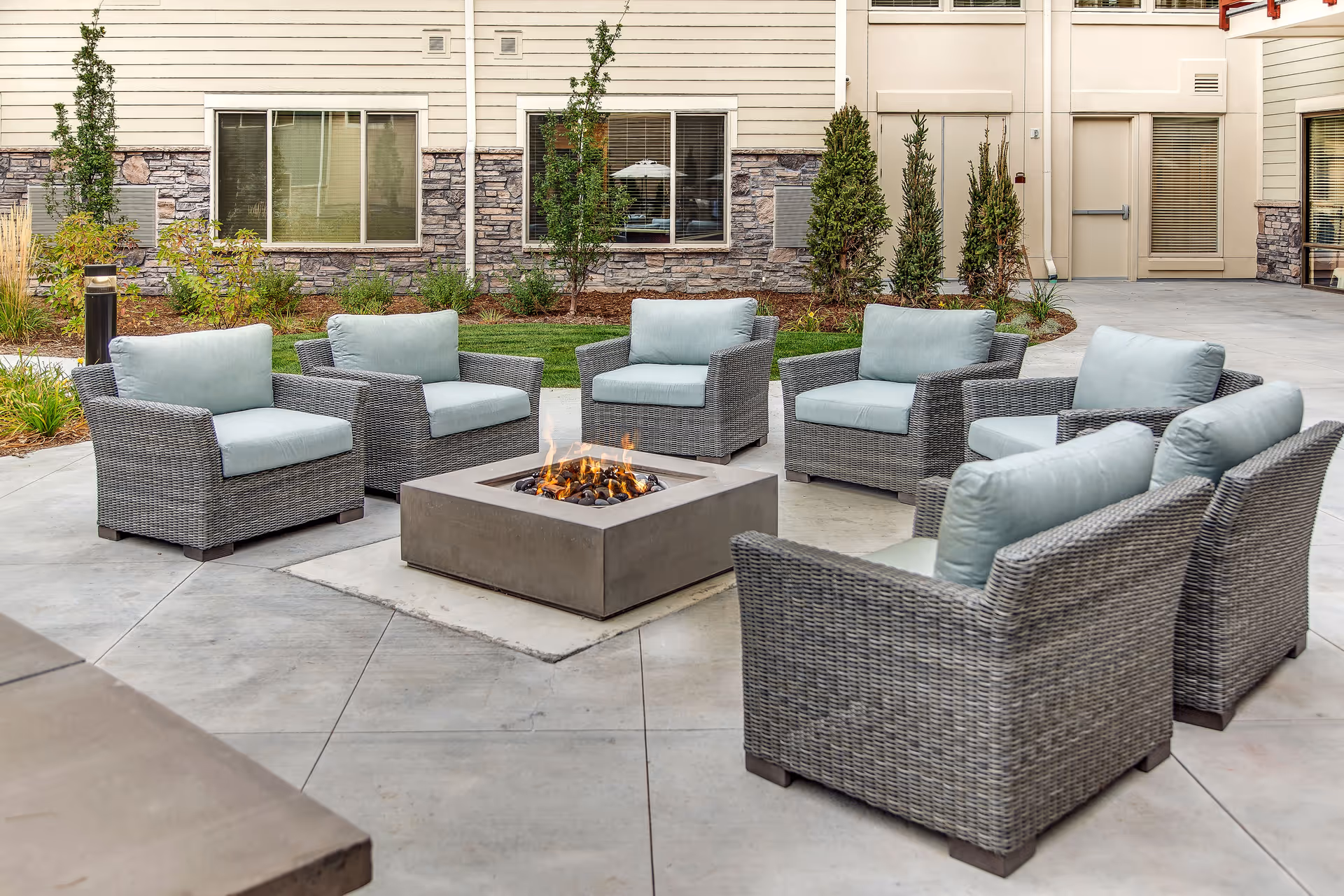 Outdoor seating area with six cushioned wicker armchairs arranged around a square fire pit with flames. The area is paved with concrete and surrounded by a building with stone and siding exterior walls, windows, and some landscaping with small trees and shrubs.