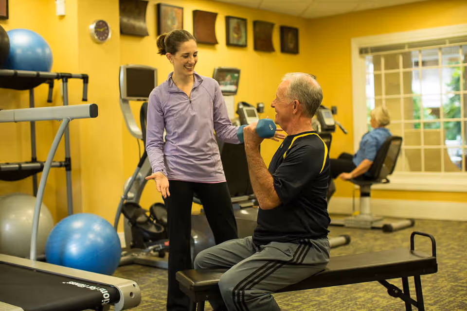 A fitness instructor assists a senior man lifting a dumbbell in a gym/exercise room.