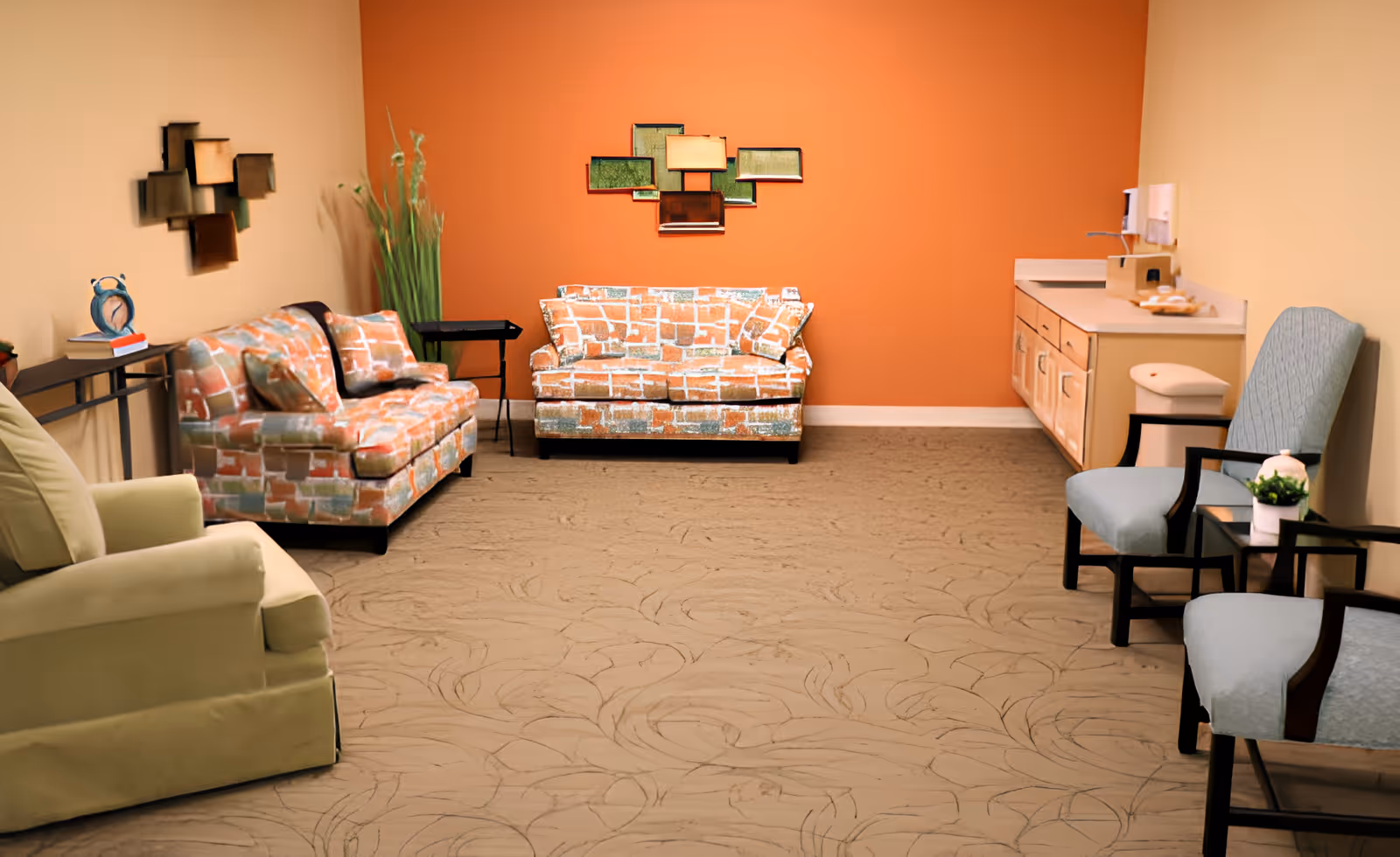 A cozy living room with patterned orange and beige sofas, a beige armchair, two blue cushioned chairs, a small black side table with a plant, and a countertop with cabinets and a sink. The walls are painted in beige and orange, with decorative wall art and tall green plants in the corner.