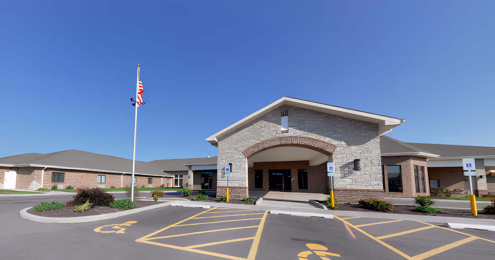 Exterior view of Brightly Senior Living facility showing the main entrance with a stone facade, a covered entryway, an American flag on a flagpole, and designated handicapped parking spaces in front.