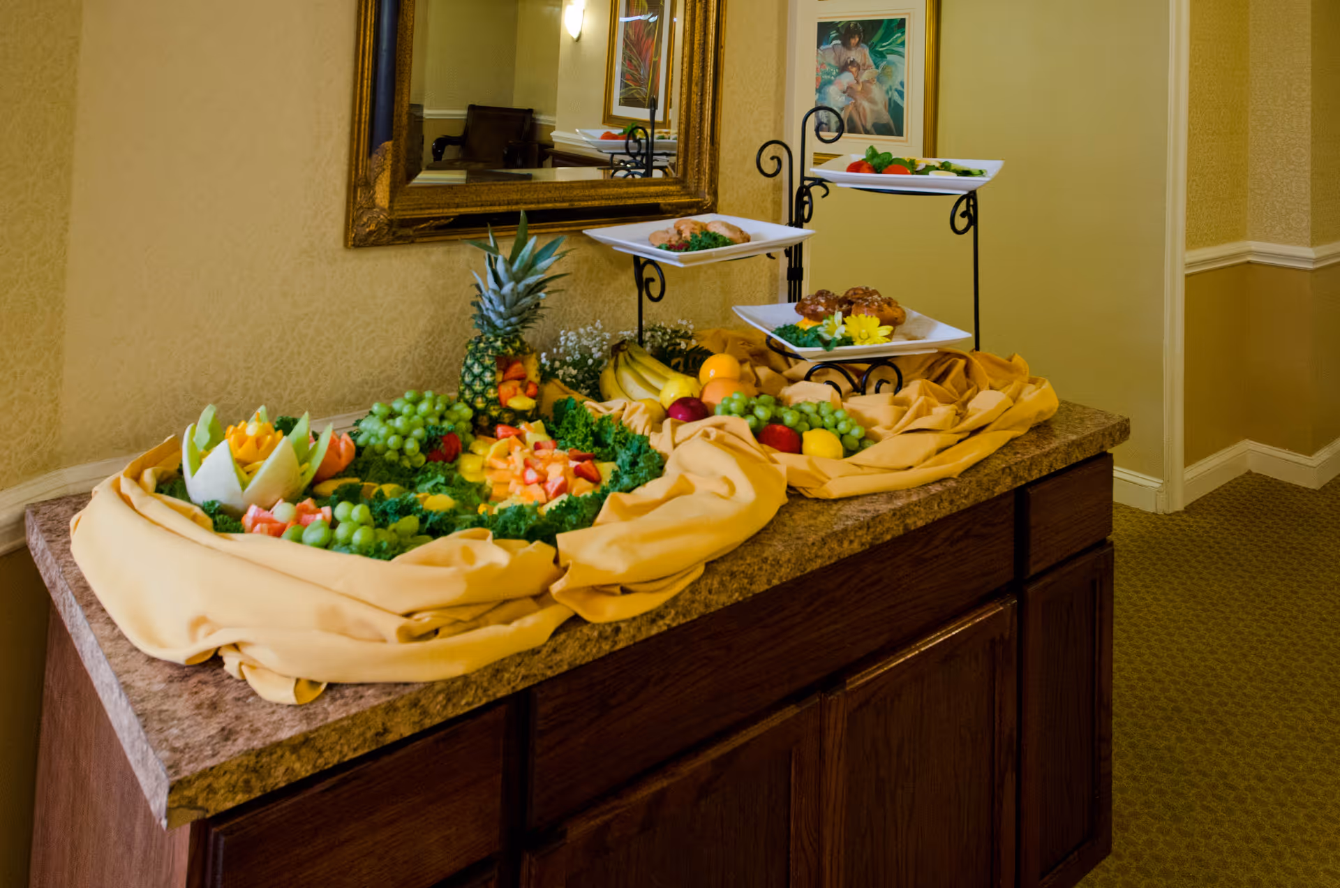 A buffet table with a variety of fresh fruits including grapes, pineapple, bananas, and melon slices arranged on a yellow cloth. There are also three white plates on a black metal stand holding different food items. The table is against a wall with a large mirror and a framed painting.