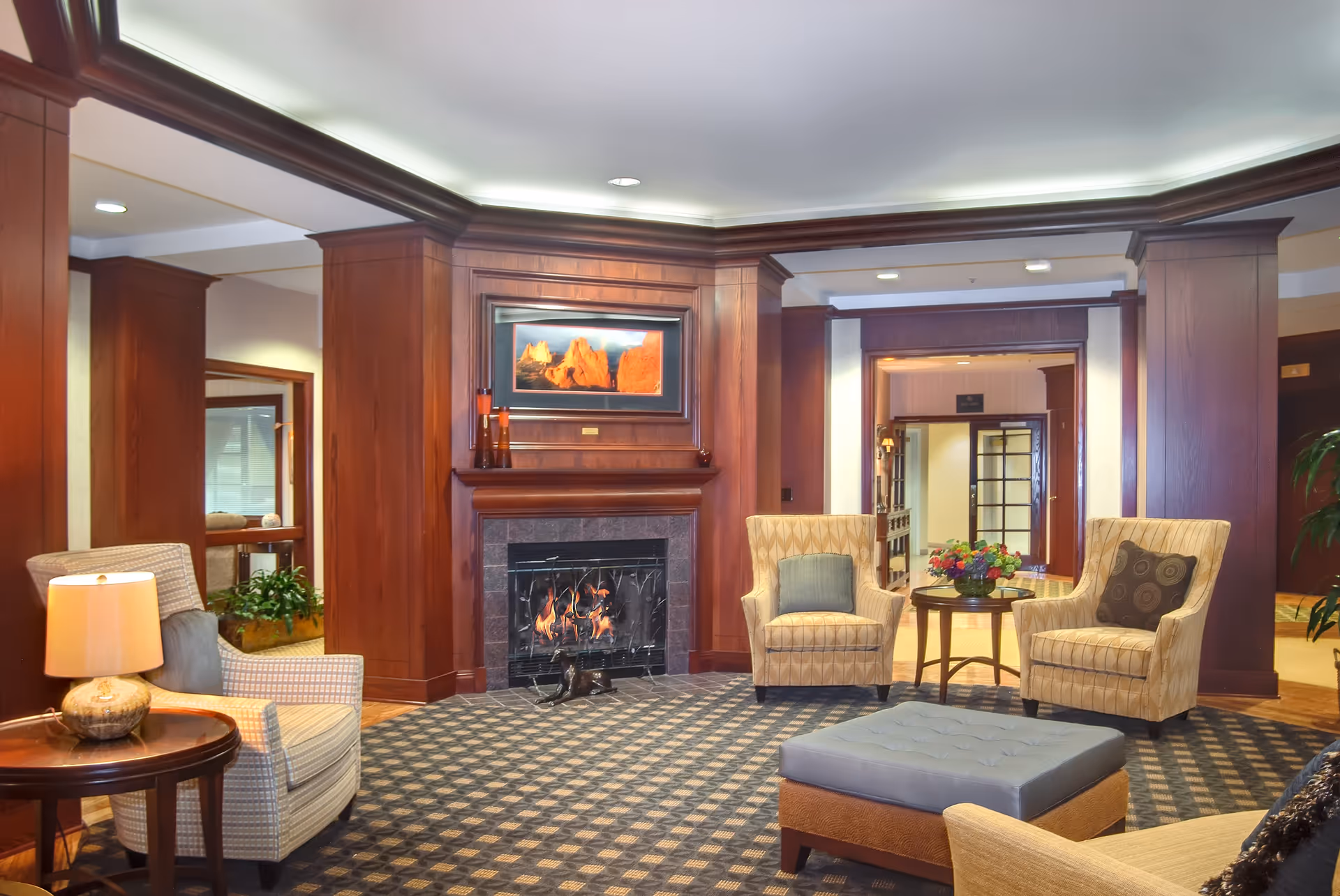 A cozy living room area with a lit fireplace surrounded by wooden paneling. The room features patterned carpet, two beige armchairs with cushions, a round wooden table with a flower arrangement, a cushioned ottoman, and a side table with a lamp. The ceiling has recessed lighting and crown molding, and there is a framed picture above the fireplace.