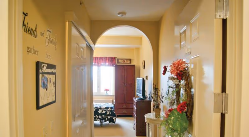 View down a hallway in a senior living facility with beige walls and an arched doorway leading to a bedroom. The hallway features decorative wall art with the words 'Friends and Family gather here' and a 'Memories' photo frame. The bedroom has a bed with a floral bedspread, a window with a red valance, a wooden armoire, and a TV on a dresser. There are also plants and floral decorations along the hallway.
