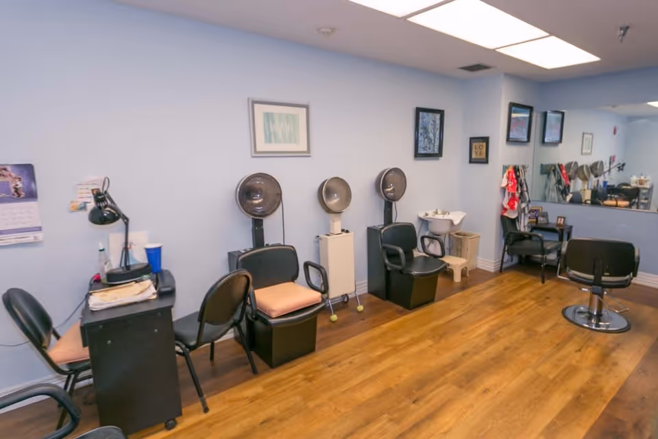 Interior view of a hair salon area with several black salon chairs, hair dryers, a small sink, a large mirror on the wall, and a few framed pictures and decorations on light blue walls. The floor is wooden.