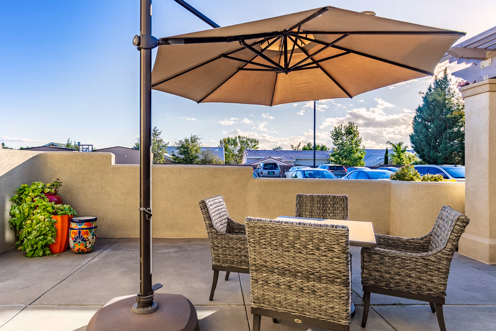 Outdoor patio area with a large beige umbrella shading a square table surrounded by four wicker chairs. There are decorative colorful pots with plants in the corner, and a low beige wall enclosing the patio. Beyond the wall, parked cars and trees are visible under a partly cloudy sky.