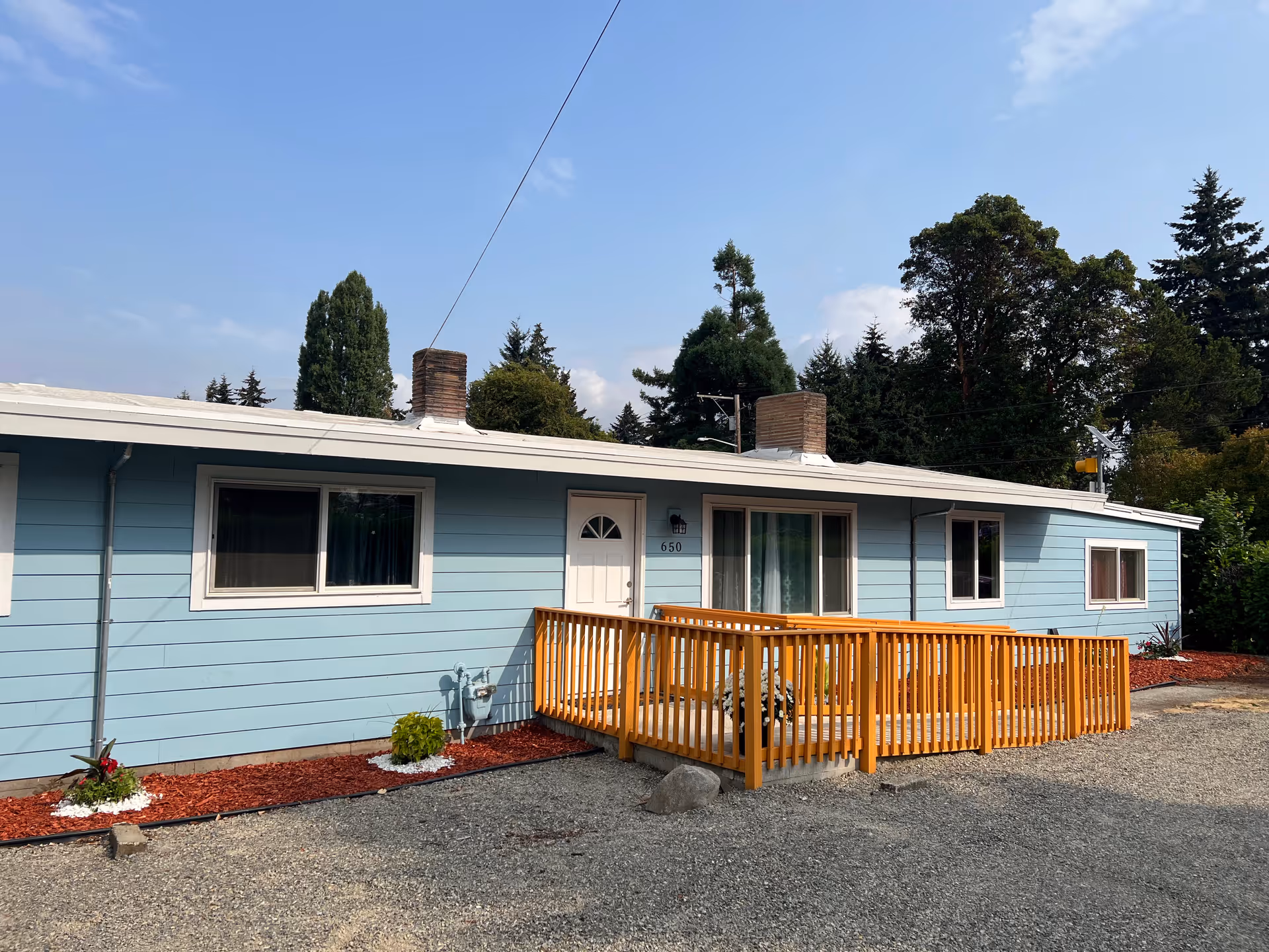 Single-story light blue building with white trim, two chimneys, a white door, and several windows. There is a wooden ramp with railings leading to the entrance. The building is surrounded by gravel and landscaped with red mulch and small plants. Trees and a clear sky are visible in the background.