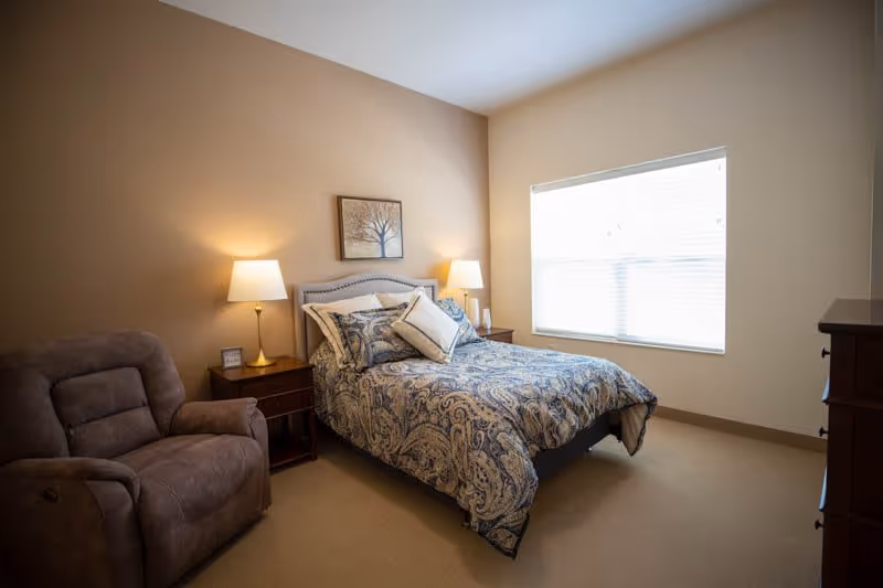 A cozy bedroom with a bed covered in a blue and white patterned comforter and pillows. There are two bedside tables with lamps on each side of the bed. A framed picture of a tree hangs on the wall above the bed. A large window with blinds lets in natural light. A comfortable brown recliner chair is positioned near one bedside table.