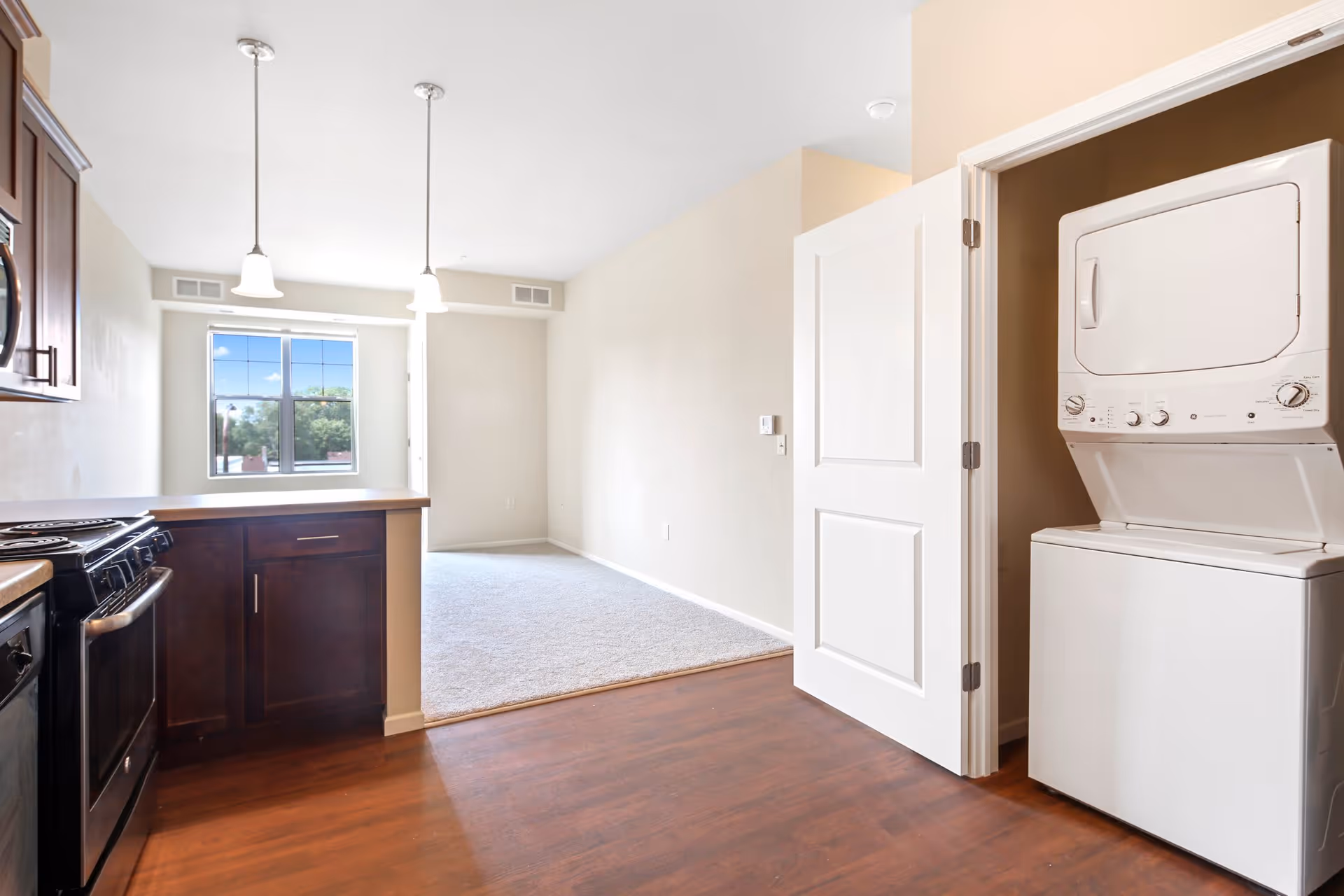 Interior view of an apartment featuring a kitchen area with dark wood cabinets, a stove, and a countertop. Adjacent to the kitchen is a carpeted living space with a window letting in natural light. On the right side, there is an open door revealing a stacked washer and dryer unit.