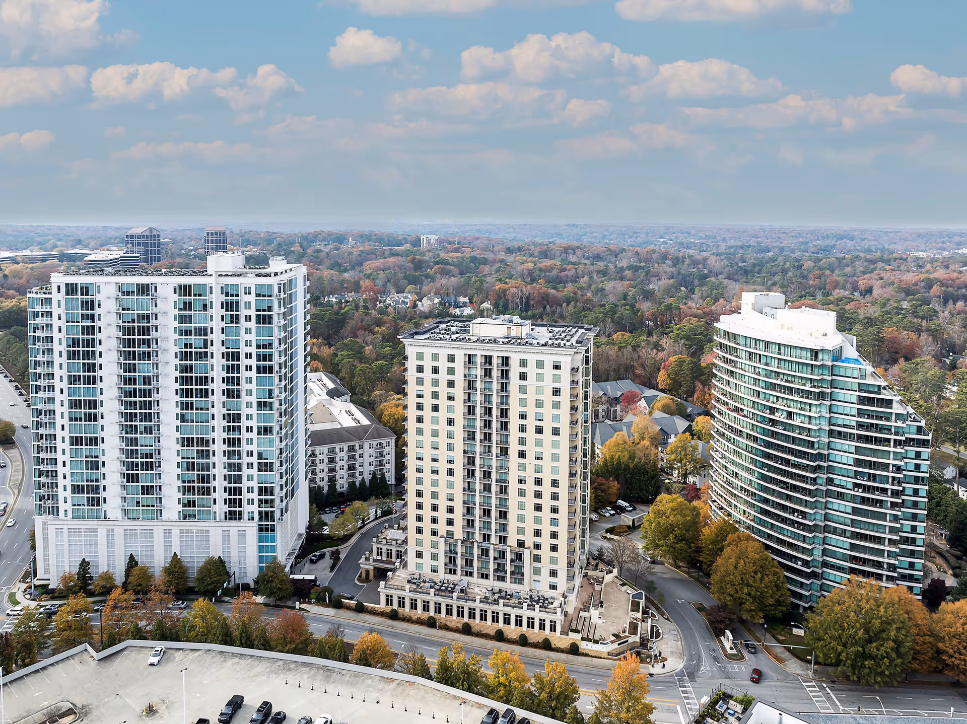 Aerial view of three high-rise residential buildings surrounded by trees and roads under a cloudy sky.
