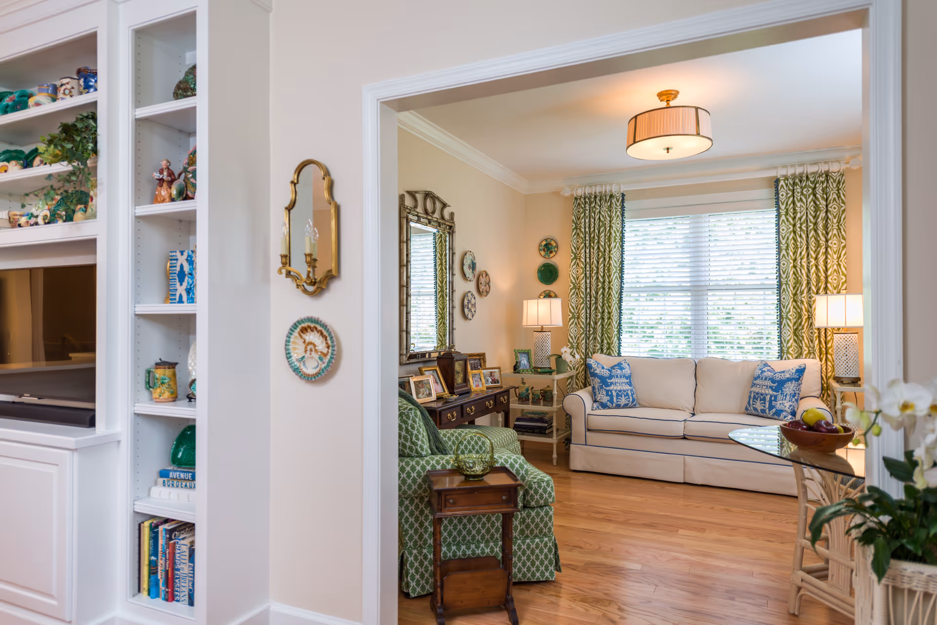 A cozy living room with a white sofa adorned with blue patterned pillows, a green armchair with a geometric pattern, wooden flooring, and large windows with green patterned curtains allowing natural light to fill the room. The room features decorative plates on the wall, a mirror, table lamps, and a glass-top table with a bowl of fruit. Adjacent to the living room is a built-in white shelving unit with various decorative items and books.