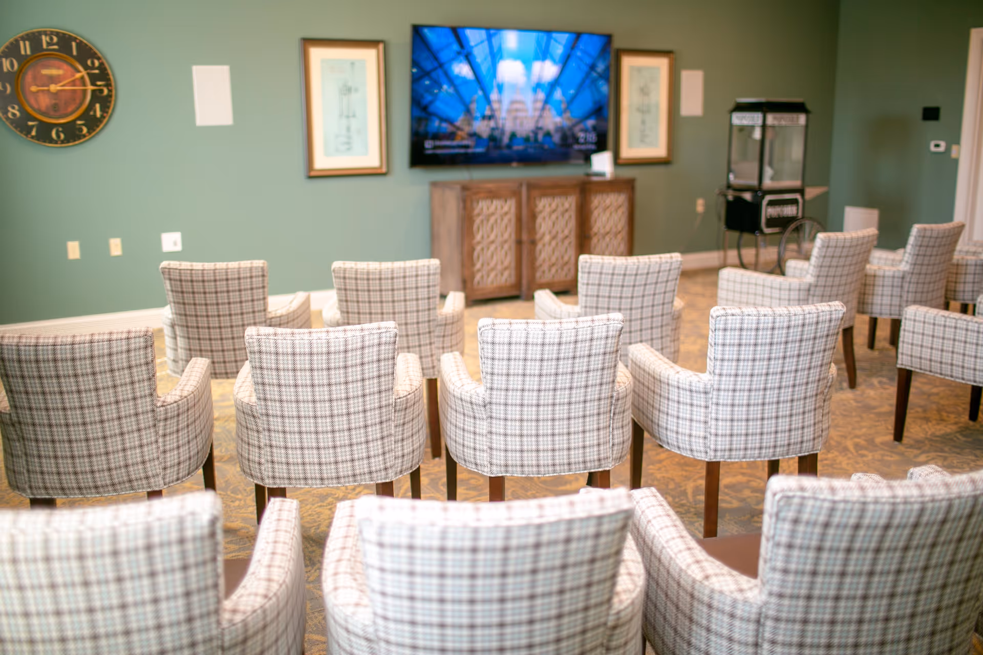 A room with multiple plaid upholstered chairs arranged in rows facing a wall-mounted flat screen TV. The wall behind the TV is painted green and decorated with two framed pictures and a large round clock. There is a wooden cabinet below the TV and a popcorn machine to the right side of the room.