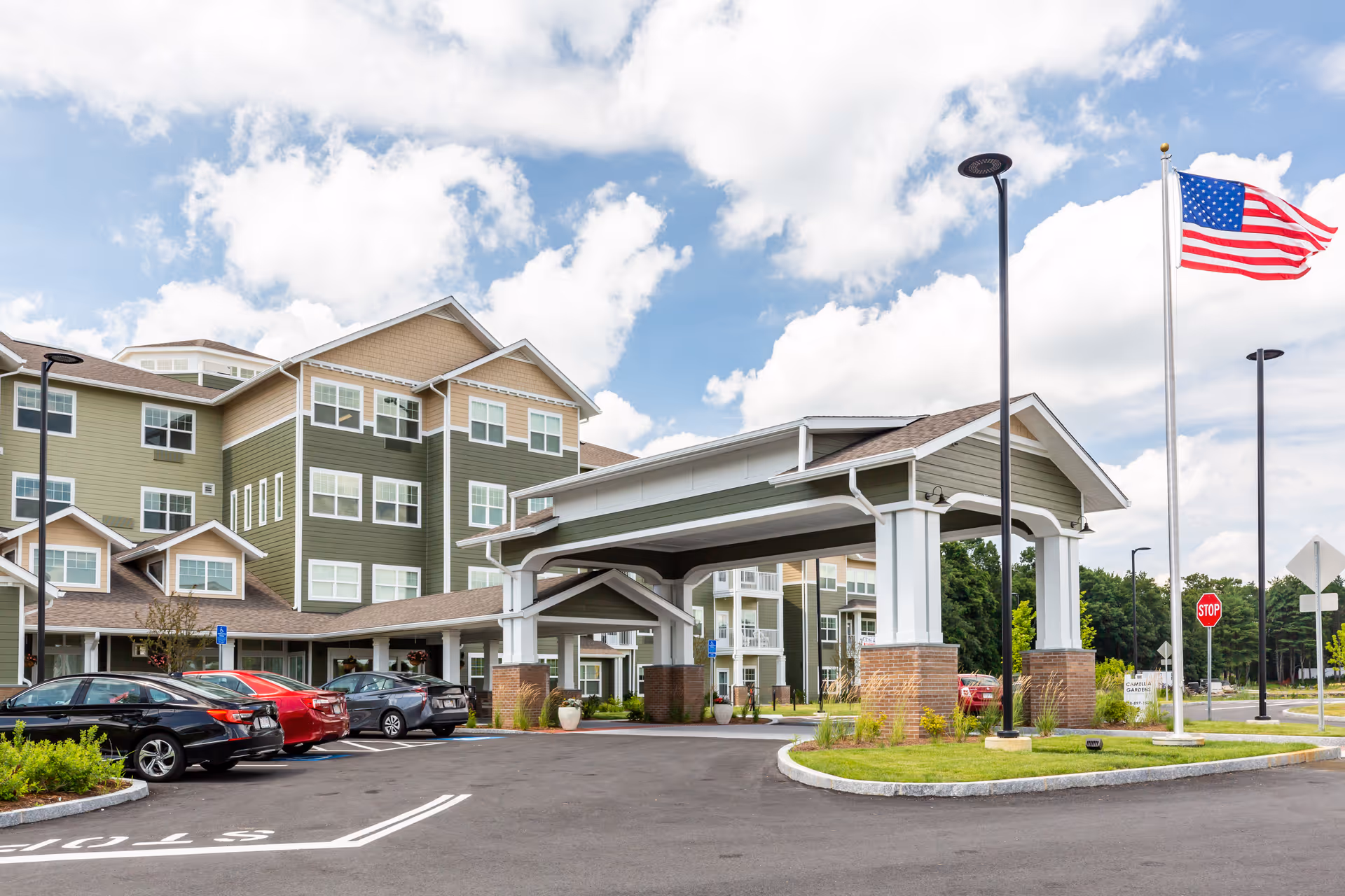 Exterior view of Camellia Gardens Gracious Retirement Living facility showing a multi-story building with green and beige siding, a covered entrance with white columns and brick bases, several parked cars, a flagpole with an American flag, and a stop sign under a partly cloudy sky.