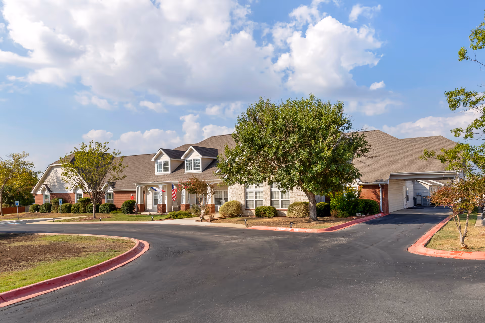Single-story senior living facility with curved driveway, manicured landscaping, and flag at the entrance.