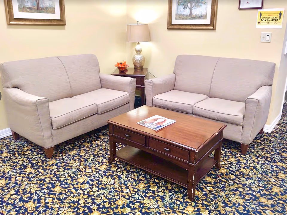 A cozy sitting area with two beige upholstered sofas facing each other, separated by a wooden coffee table with magazines on top. There is a side table with a decorative lamp and a small flower arrangement between the sofas. The room has patterned carpet flooring and framed artwork on the walls.