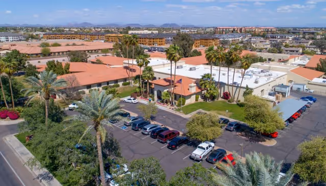 Aerial view of Peoria Post Acute facility showing multiple buildings with red-tiled roofs, surrounded by palm trees and greenery. There is a parking lot with several cars parked, and the background includes a sprawling urban area under a partly cloudy sky.