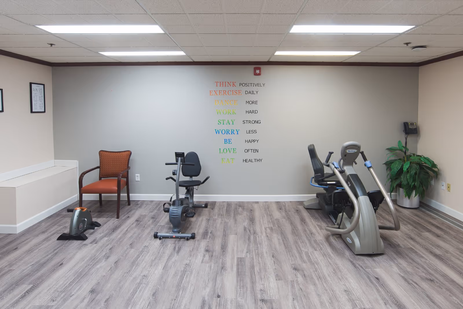 A small exercise room with light wood flooring and beige walls. The room contains two stationary exercise bikes and one recumbent exercise bike. There is a single orange cushioned chair on the left side and a green potted plant on the right side. On the back wall, colorful motivational words are displayed vertically: THINK, EXERCISE, DANCE, WORK, STAY, WORRY, BE, LOVE, EAT, each followed by a positive phrase such as 'POSITIVELY', 'DAILY', 'MORE', 'HARD', 'STRONG', 'LESS', 'HAPPY', 'OFTEN', and 'HEALTHY'.