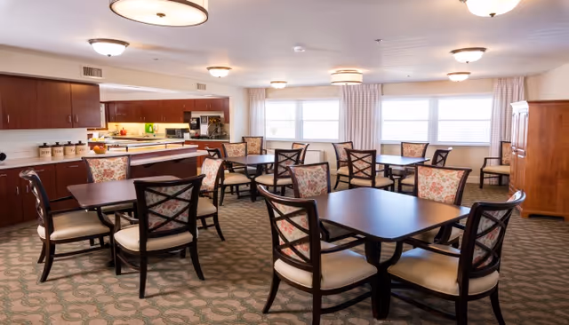 A bright dining room with several dark wood tables and chairs featuring floral patterned cushions. The room has large windows with light curtains, a carpeted floor with a patterned design, and a kitchen area with wooden cabinets and countertops in the background.