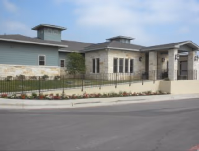 Exterior view of a single-story building with stone and light blue siding, featuring multiple windows, a covered entrance, a black metal fence, and a flower bed along the sidewalk under a partly cloudy sky.