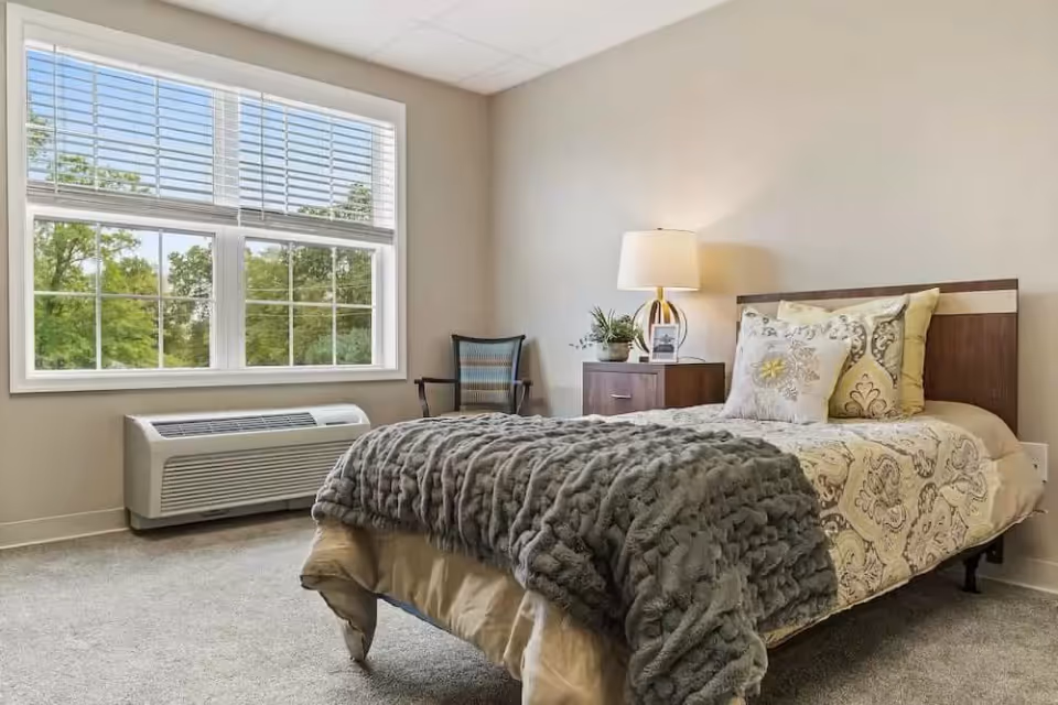 A cozy bedroom with a single bed covered in a patterned bedspread and a gray textured blanket. There are three decorative pillows on the bed. Next to the bed is a wooden nightstand with a lamp and a small plant. A chair is placed against the wall near a large window with white blinds, showing green trees outside. The room has beige walls and carpeted floor.