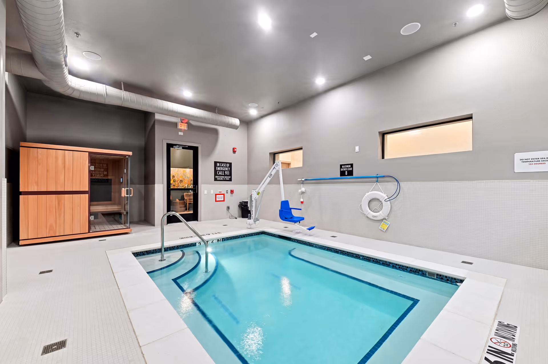 Indoor pool area with a small rectangular pool, a metal handrail, a blue pool lift chair, and a wooden sauna cabin in the corner. The room has gray walls, two small windows, and ceiling lights. Safety signs and a life preserver are mounted on the walls.