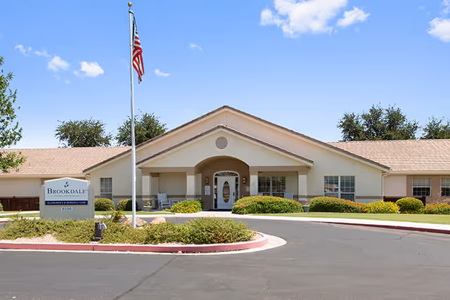 Exterior front view of Brookdale Union Hills senior living facility with a single-story building, an American flag on a flagpole, and landscaped bushes and trees under a blue sky with some clouds.